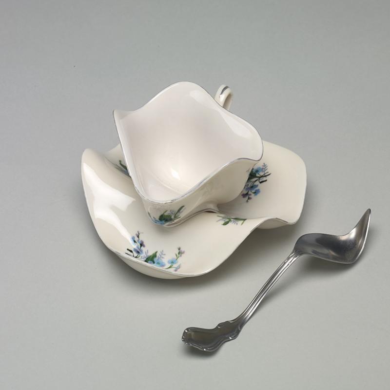 Sculptural artwork depicting a distorted white teacup and saucer with a warped silver spoon. The ceramic forms appear soft and melted, bending in an unnatural way that challenges the functional shape of the objects. Set against a plain gray background, the piece evokes surrealism and transformation
