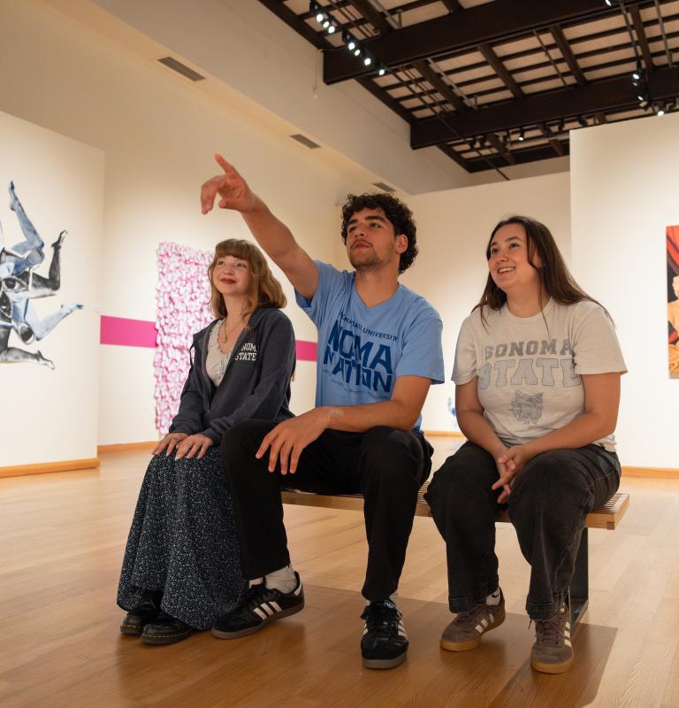 Three Sonoma State University students sit on a bench in an art gallery, smiling as one points toward artwork displayed on the walls. The gallery features large contemporary paintings and sculptures in a bright, open exhibition space.