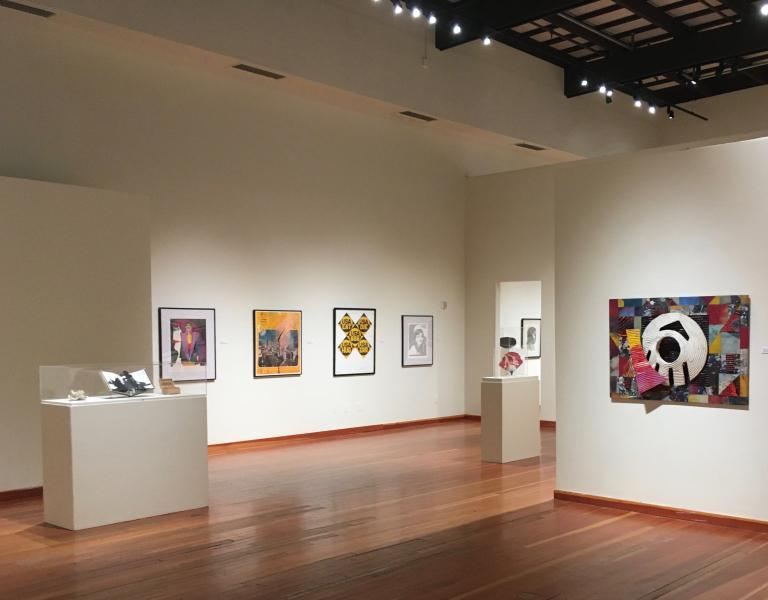 Interior view of an art gallery with white walls and wooden floors, displaying a variety of contemporary artworks. Framed paintings and photographs hang on the walls, while sculptures are presented on white pedestals. The room is brightly lit with ceiling track lights, creating a clean and modern exhibition space
