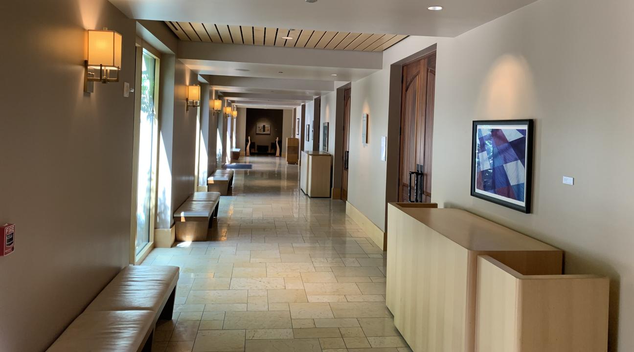 Interior view of a well-lit art gallery hallway with cream-colored walls and carpeted flooring. Framed artworks hang along the walls, including a blue-toned abstract piece on the right. Warm wall sconces provide ambient lighting, and natural light streams in from windows on the left. A seating area is visible at the far end of the corridor