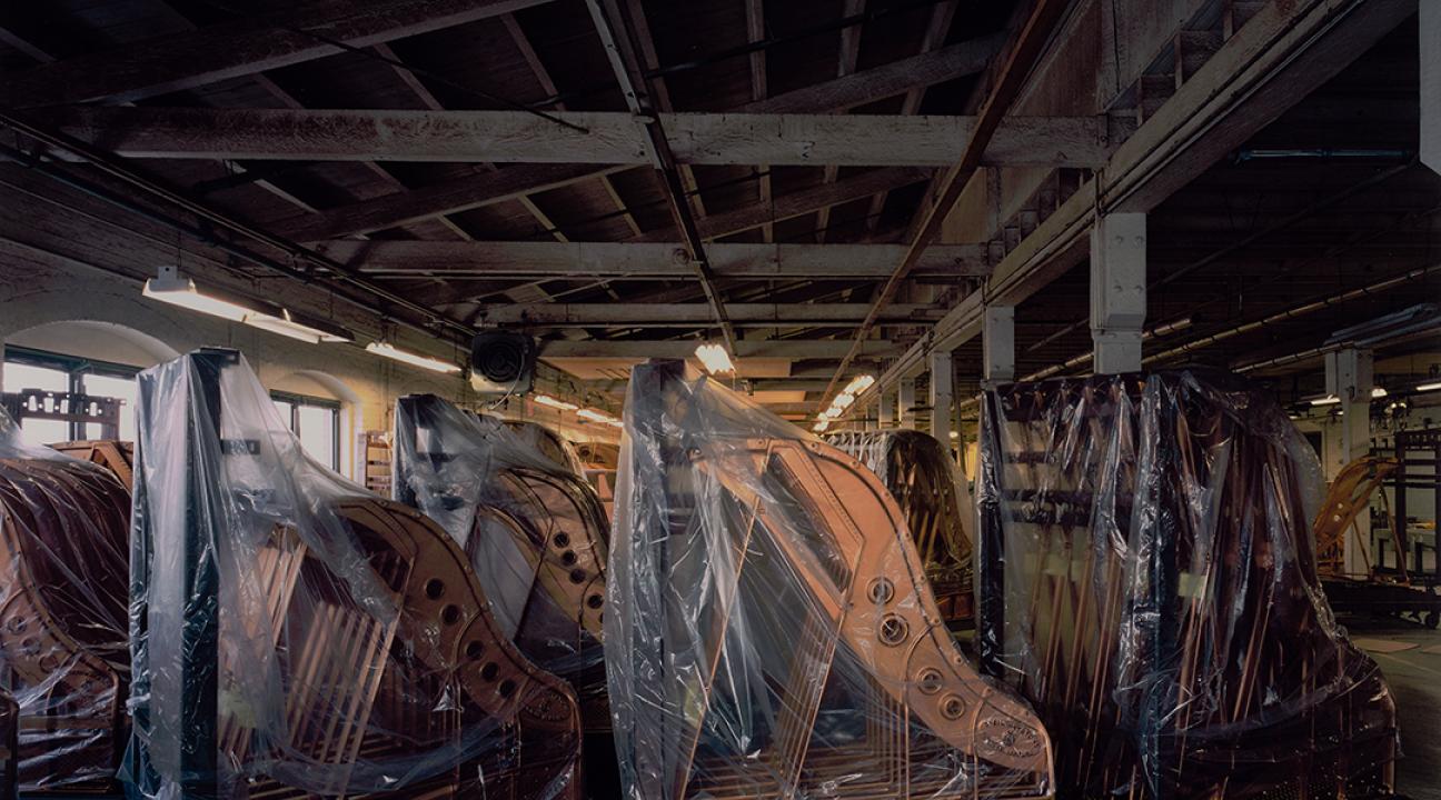 Photograph of an old, dimly lit warehouse filled with rows of grand pianos, many covered in protective plastic sheets. The wooden beams and exposed ceiling give the space a weathered, industrial feel. Light from the right softly illuminates the instruments, creating a quiet, melancholic atmosphere