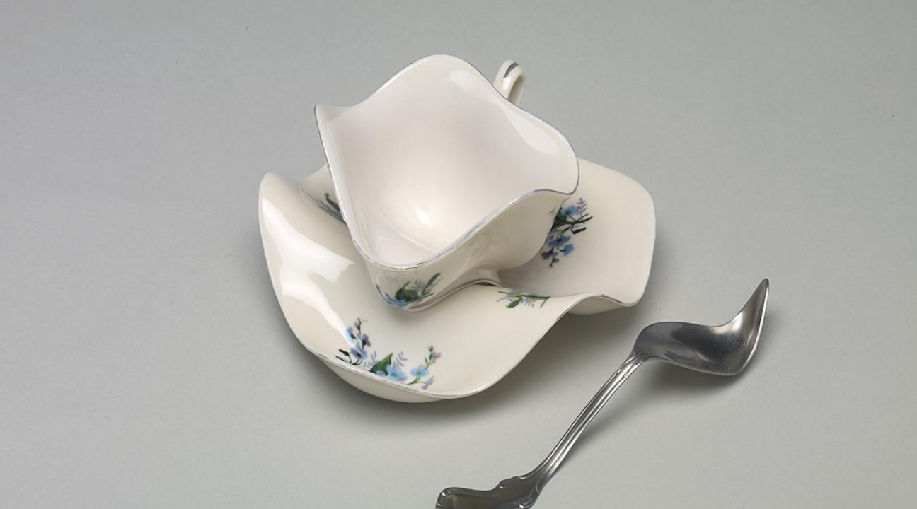 Sculptural artwork depicting a distorted white teacup and saucer with a warped silver spoon. The ceramic forms appear soft and melted, bending in an unnatural way that challenges the functional shape of the objects. Set against a plain gray background, the piece evokes surrealism and transformation