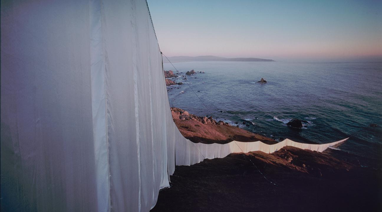 Photograph showing a massive white fabric installation draped over coastal cliffs, extending toward the ocean at sunset. The fabric billows slightly in the wind, contrasting against the rugged landscape and calm sea below. The image captures the monumental and ephemeral nature of environmental art.