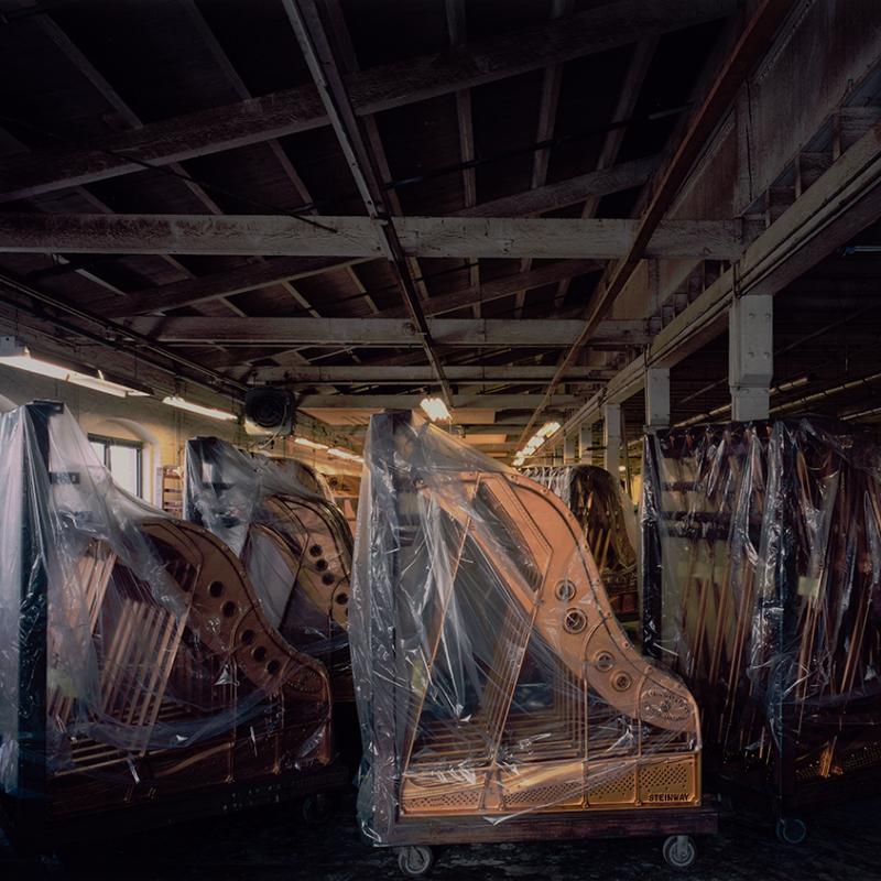 Photograph of an old, dimly lit warehouse filled with rows of grand pianos, many covered in protective plastic sheets. The wooden beams and exposed ceiling give the space a weathered, industrial feel. Light from the right softly illuminates the instruments, creating a quiet, melancholic atmosphere