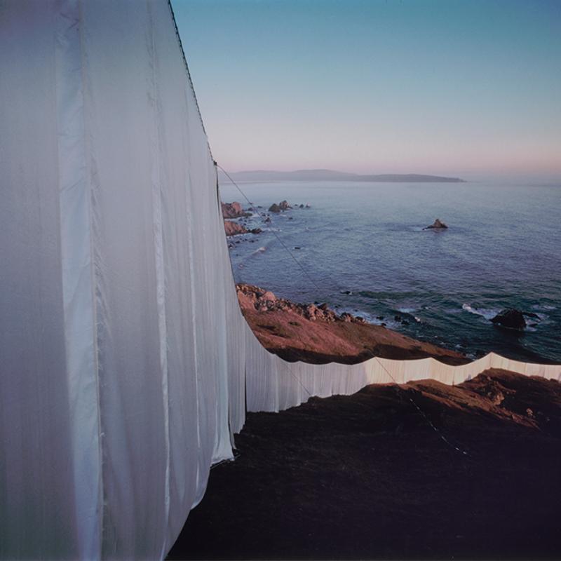 Photograph showing a massive white fabric installation draped over coastal cliffs, extending toward the ocean at sunset. The fabric billows slightly in the wind, contrasting against the rugged landscape and calm sea below. The image captures the monumental and ephemeral nature of environmental art.