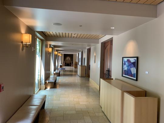 Interior view of a well-lit art gallery hallway with cream-colored walls and carpeted flooring. Framed artworks hang along the walls, including a blue-toned abstract piece on the right. Warm wall sconces provide ambient lighting, and natural light streams in from windows on the left. A seating area is visible at the far end of the corridor