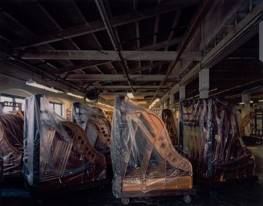 Photograph of an old, dimly lit warehouse filled with rows of grand pianos, many covered in protective plastic sheets. The wooden beams and exposed ceiling give the space a weathered, industrial feel. Light from the right softly illuminates the instruments, creating a quiet, melancholic atmosphere