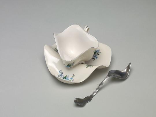 Sculptural artwork depicting a distorted white teacup and saucer with a warped silver spoon. The ceramic forms appear soft and melted, bending in an unnatural way that challenges the functional shape of the objects. Set against a plain gray background, the piece evokes surrealism and transformation
