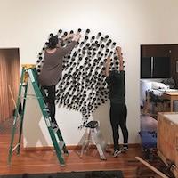 Two people install an art piece on a gallery wall. The artwork consists of numerous small black objects arranged in a circular, clustered pattern. One person stands on a ladder while the other adjusts pieces from the floor. The room has wooden floors and white walls, with open doorways visible on either side