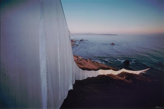 Photograph showing a massive white fabric installation draped over coastal cliffs, extending toward the ocean at sunset. The fabric billows slightly in the wind, contrasting against the rugged landscape and calm sea below. The image captures the monumental and ephemeral nature of environmental art.